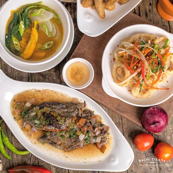 A bowl of sinigang, sinaing na tulingan on an oval dish, and salt and pepper squid rings on a wooden table, 2023