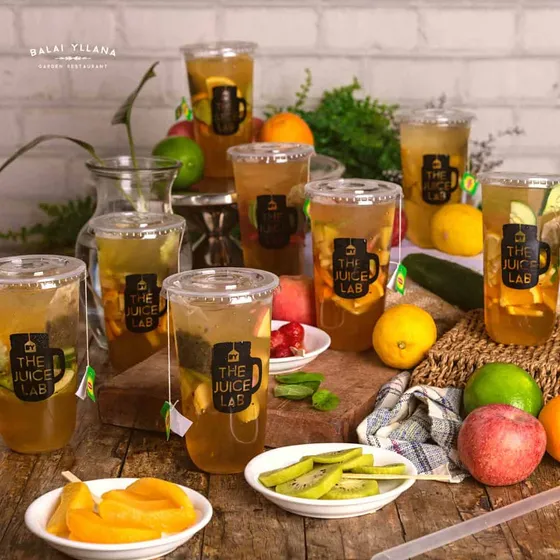 Cups of juice blends with sliced fruit, labelled The Juice Lab, with lemons, limes, apples and kiwi on a wooden table, 2023