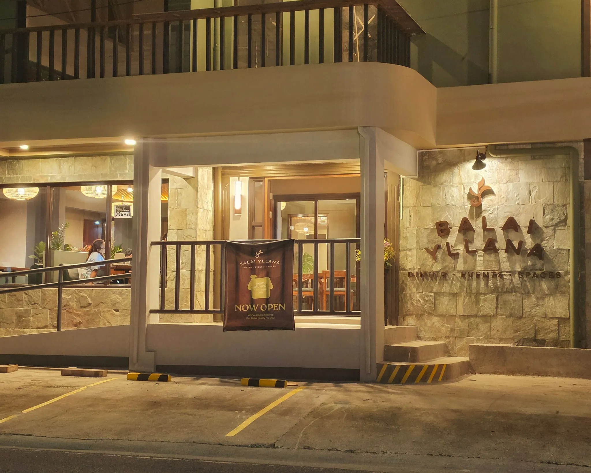 The front of Balai Yllana at dusk: a stone wall with the logo beside the entrance, the dining room lit behind the windows